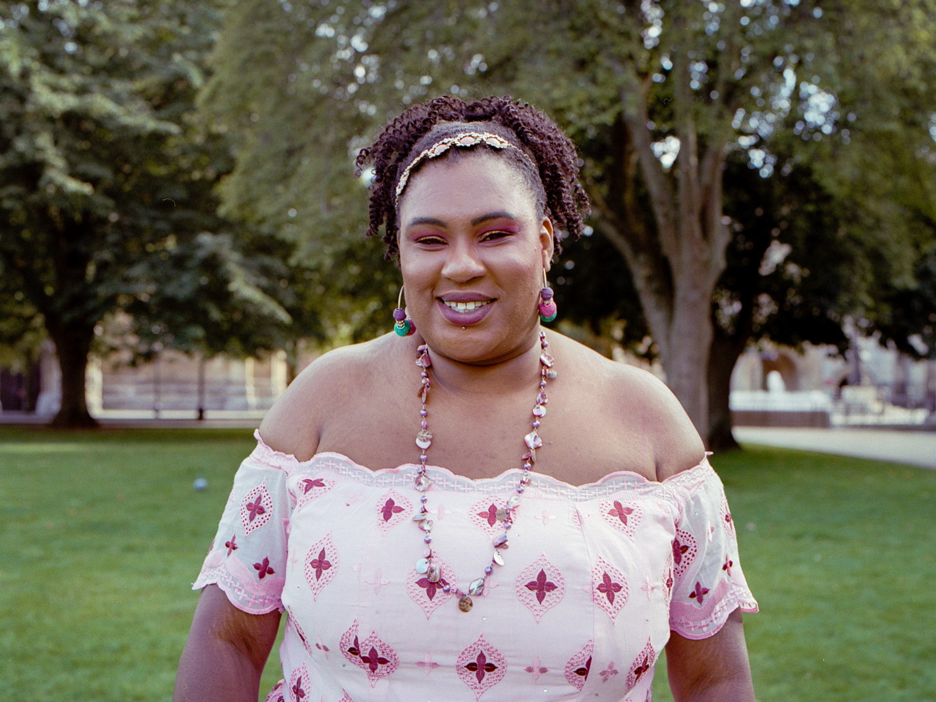 El is standing in a park wearing a pink dress. She is smiling into the camera.