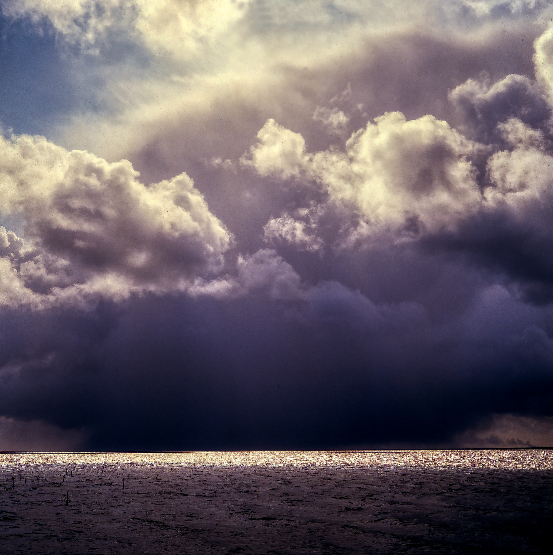 Sea and large ominous clouds with rain clouds far away. The sun shines a sliver of sunlight on the sea
