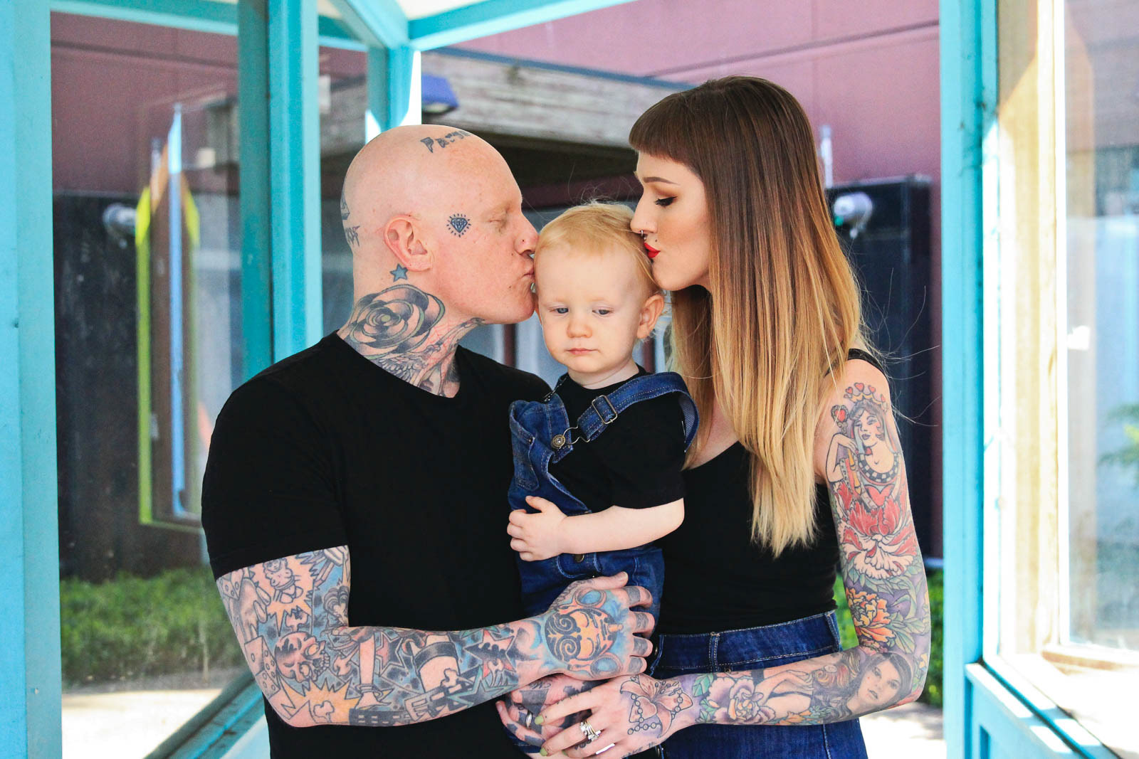 Tattooed mother and father holding young son and kissing his head