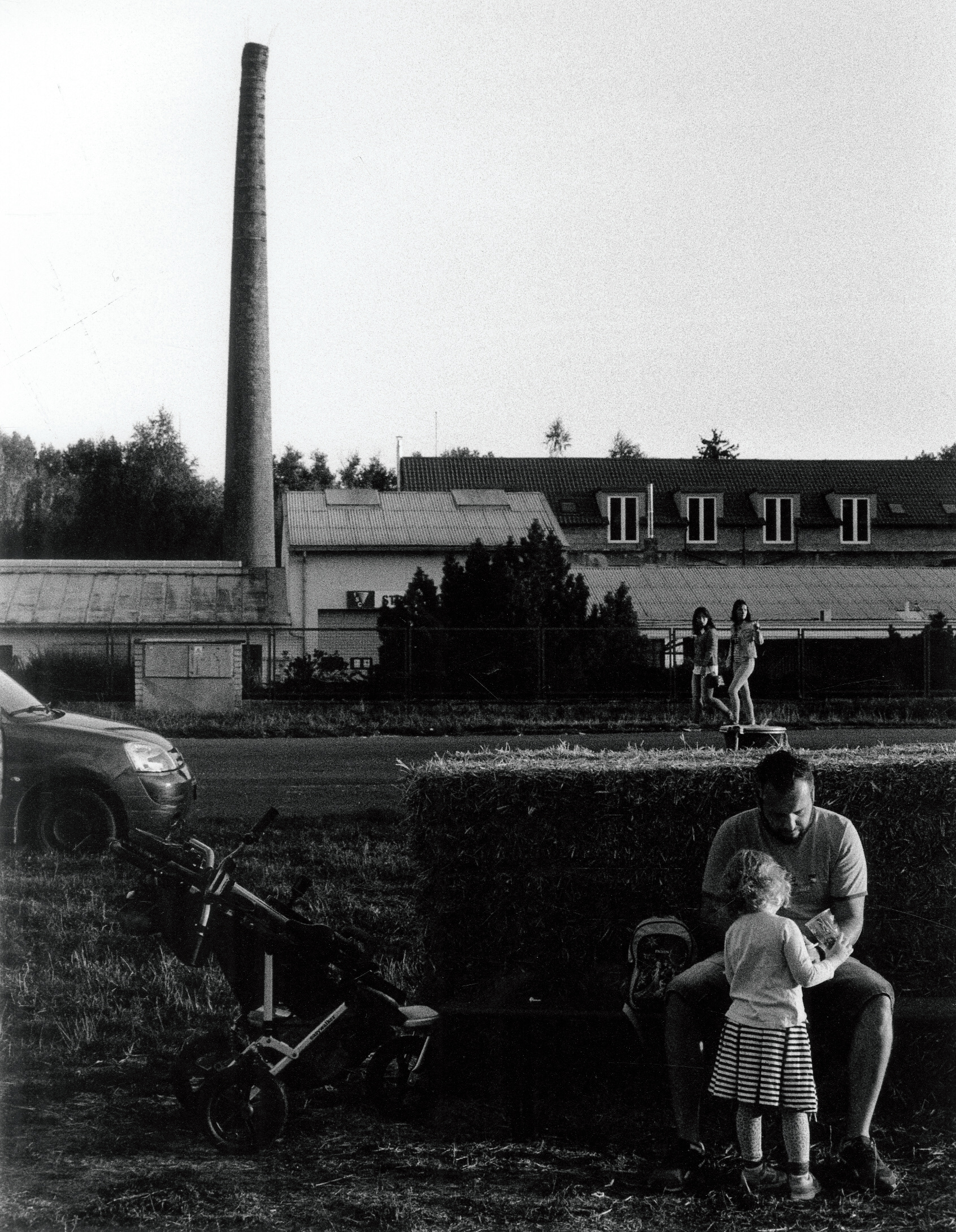 a man dotes on his daughter while sat on a haybale in a park, in the background, an old chimney stack is visible