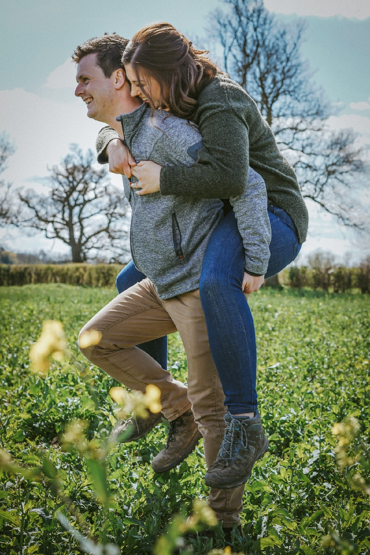 couple in piggy back smiling happily in rapeseed field on sunny day at pre wedding engagement shoot with david lefebvre a wedding photographer covering buckinghamshire and milton keynes