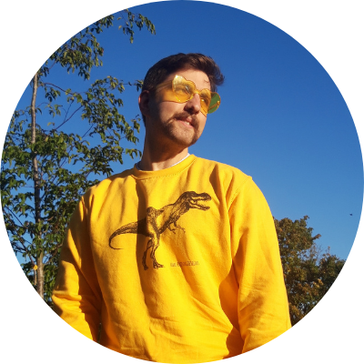 Bristol-based musician, writer and cultural producer, Chris Vox, enjoying the sun. Chris is dressed in a t-rex-themed yellow jumper and yellow heart-shaped sunglasses. He is standing in a park with bright blue sky above.