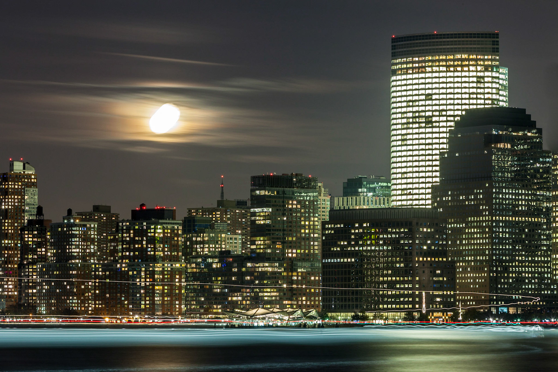 A view of the moon rising between clouds over Downtown Manhattan. A Hudson River ferry lit up the foreground while manoevering in a wide turn in front of the camera.