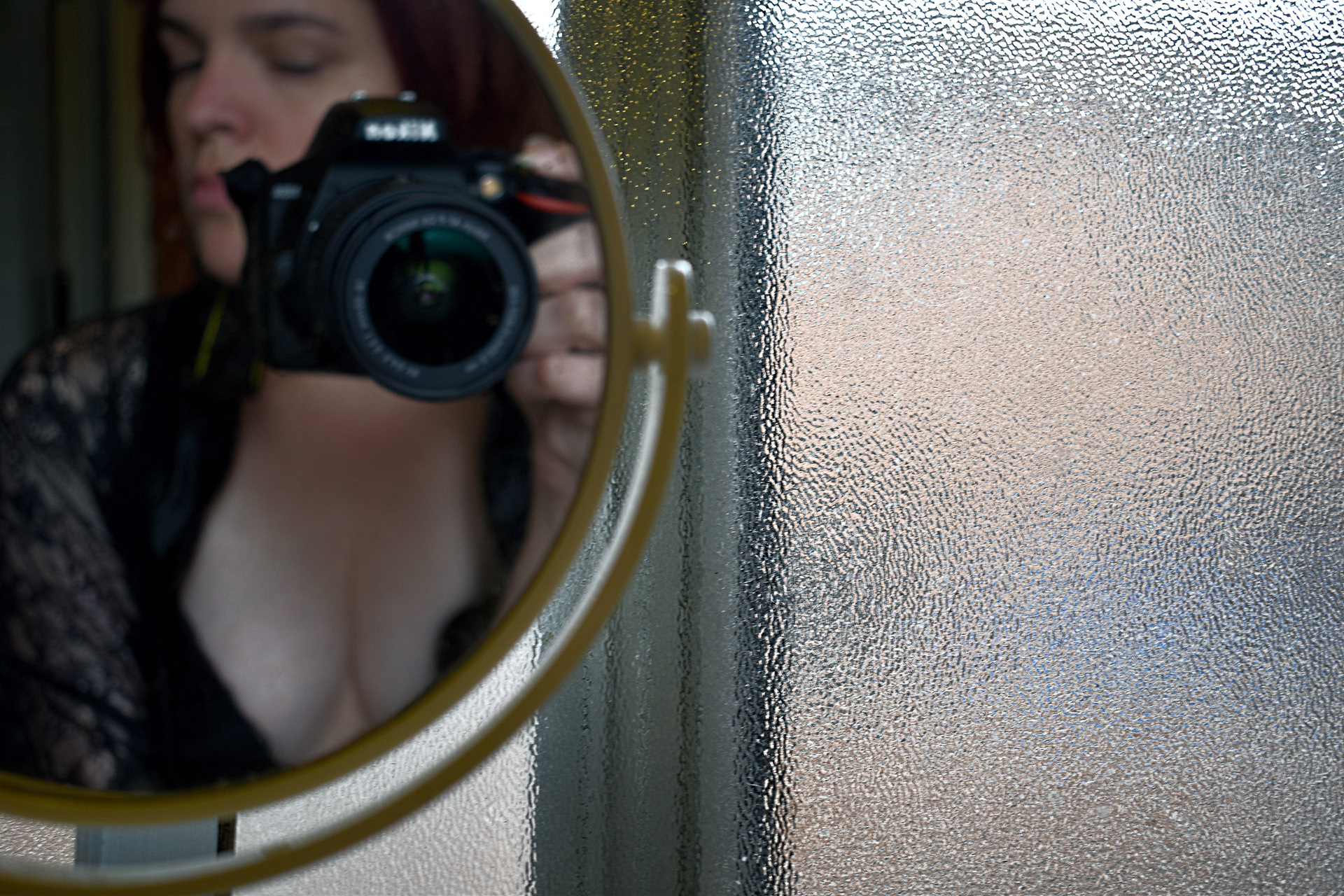 A self-portrait in a bathroom mirror, near a window, with a camera in hand. She is wearing a black negligee