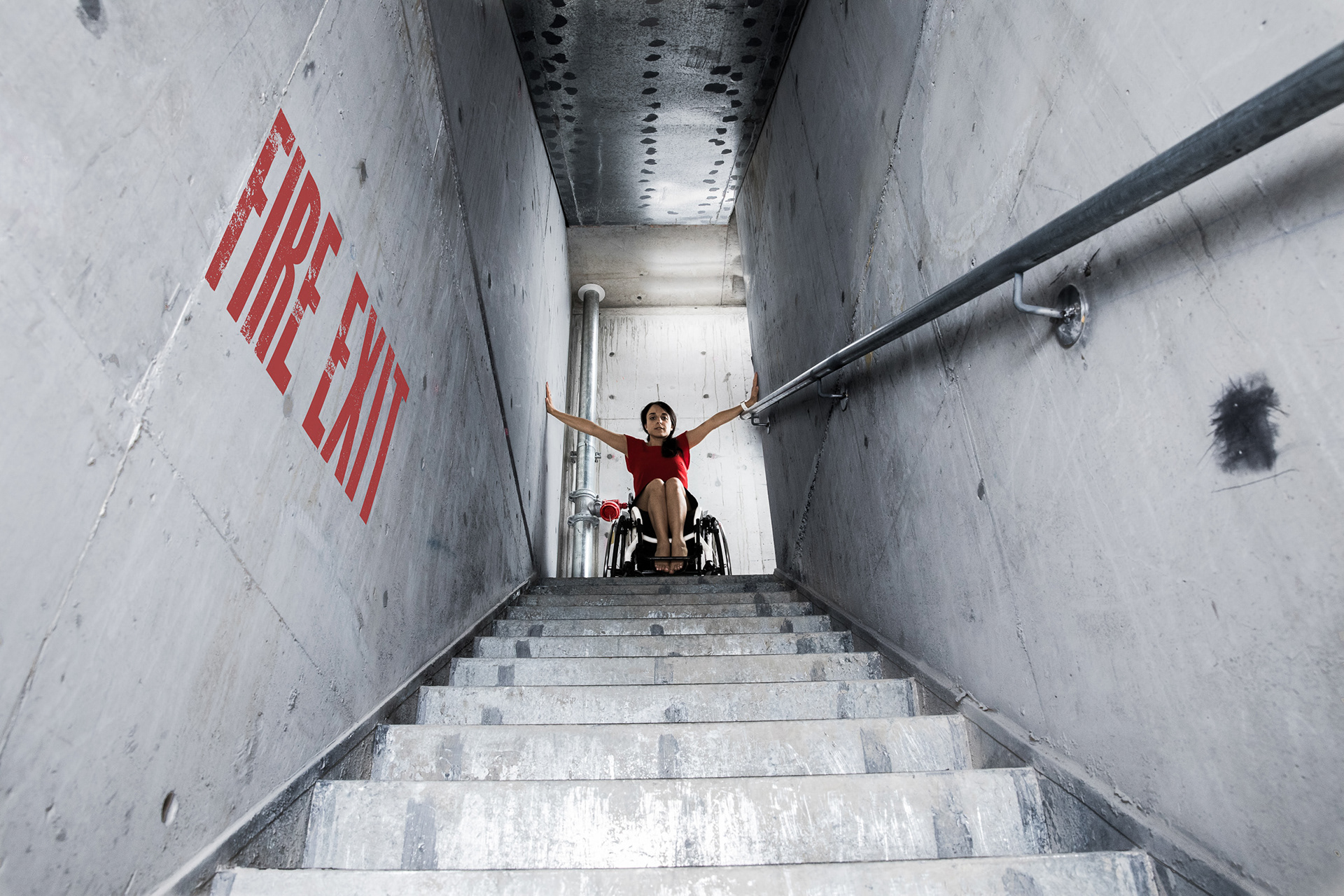 Evi in her wheelchair at the top of fire stairs looking down a grey stairwell