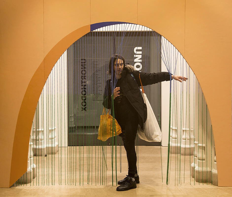 An interration of "Paradise of Greenery" at the Jewish Museum in New York. The sculpture is an archway at the entry to the gallery space, constructed out of pink foam with a beaded curtain underneath. A viewer stands underneath the archway while pulling the beaded curtain open with his arms to the right of the image.