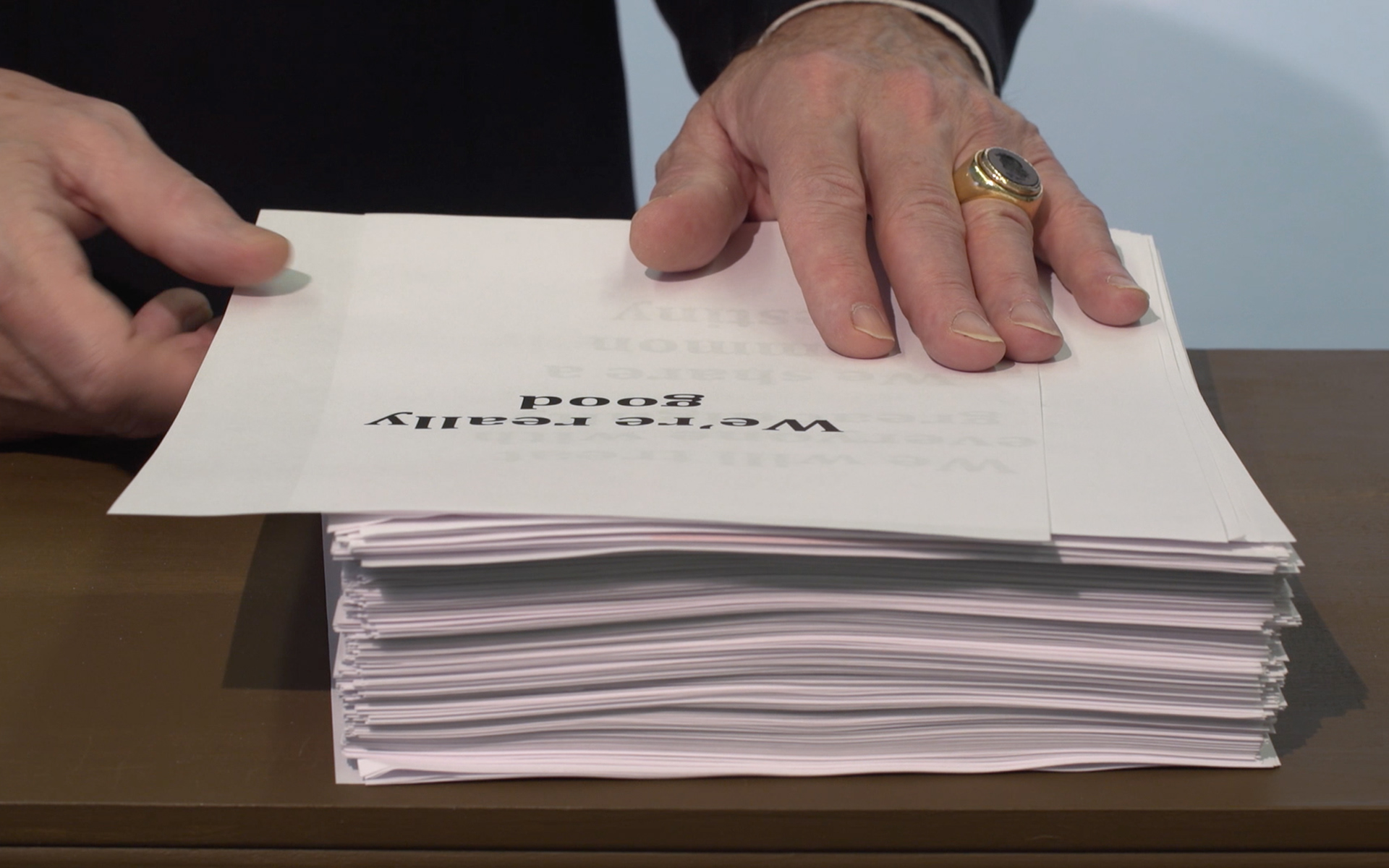 An image still from the short film "Man Above the Fog." The still shows a close-up of the man's hand resting on the large stack of papers. the top page reads: "We're really good" in large font the takes up most of the page.